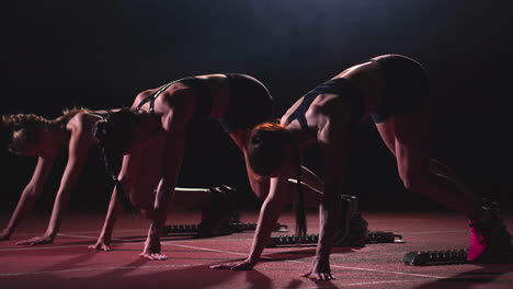 Three-girls-in-black-clothes-are-in-the-starting-pads-to-start-the-race-in-the-competition-in-the-light-of-the-lights-and-run-towards-the-finish