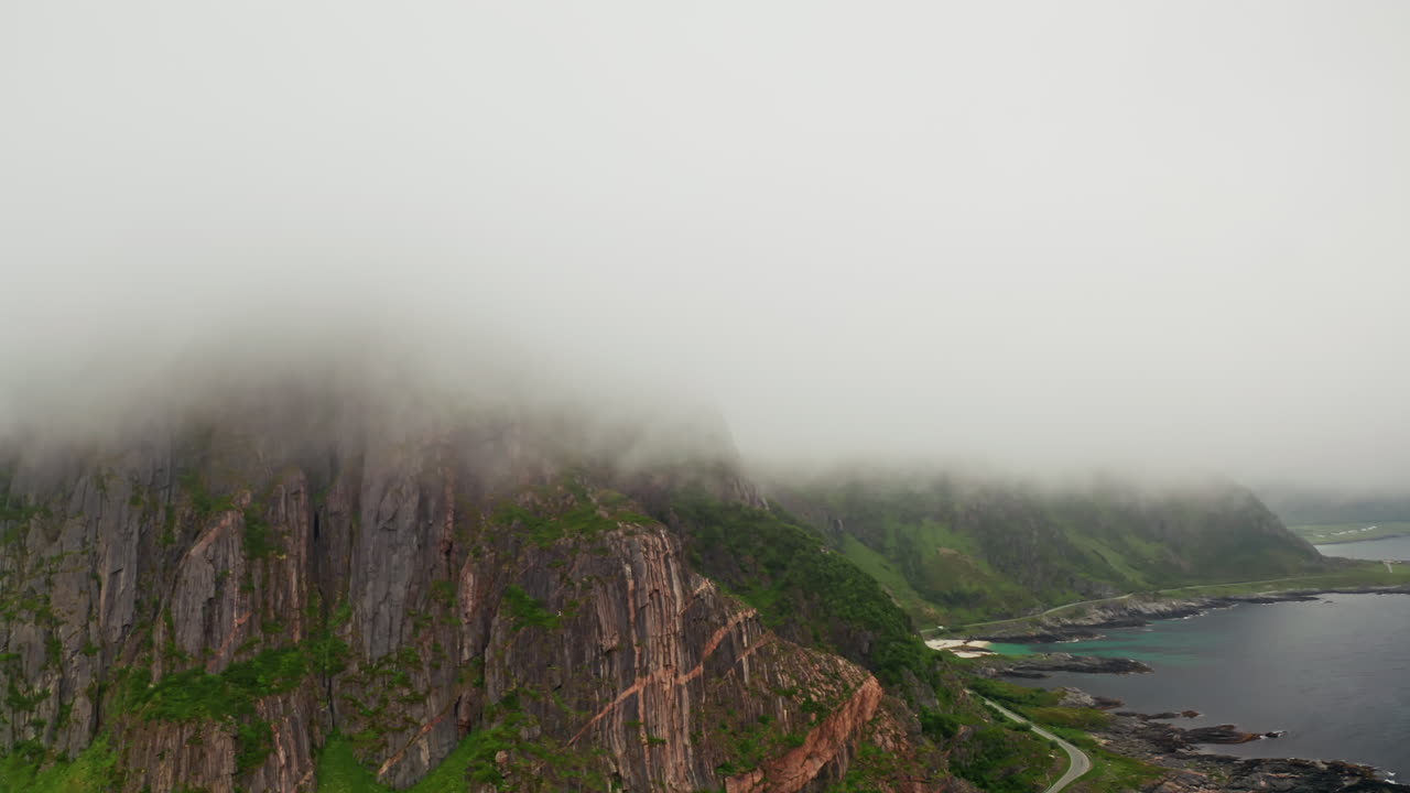 Aerial drone shot over the misty coastline of the island of Senja, Lofoten, Norway. High view of the green landscape, the sea, the rocky coastline and dramatic misty clouds coverming the mountains.