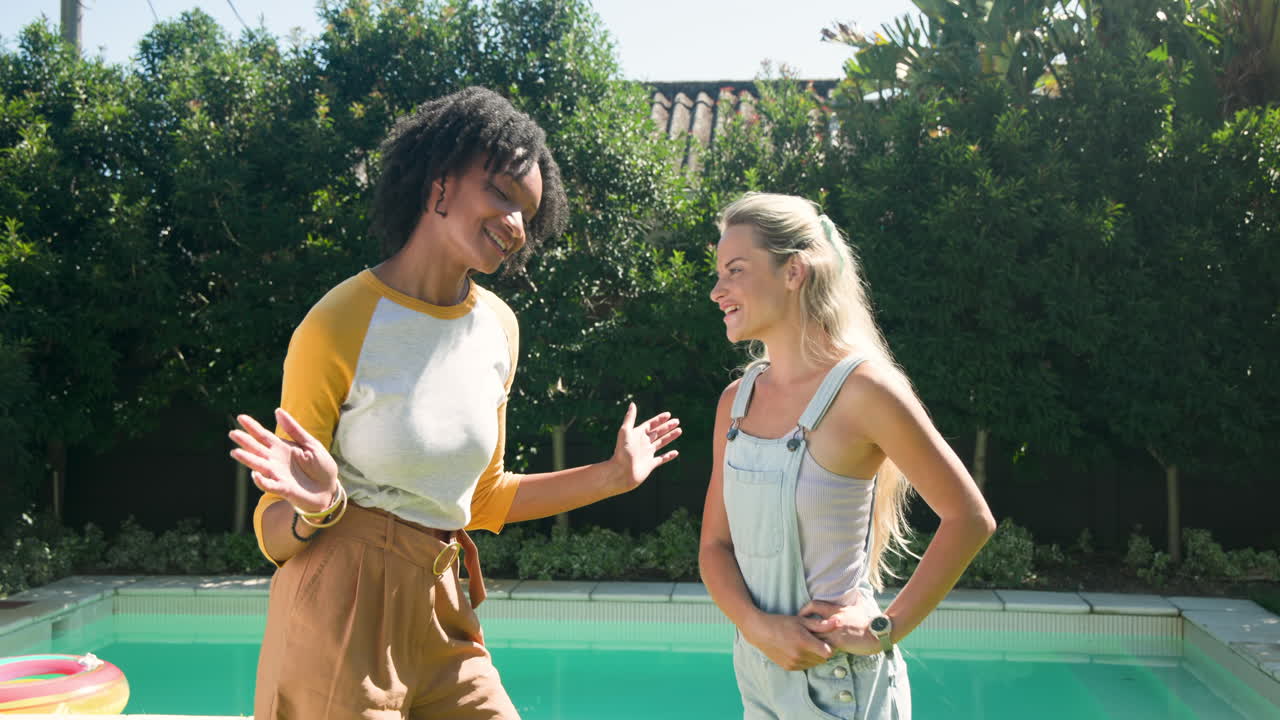 Two female friends chatting and laughing by pool on sunny day