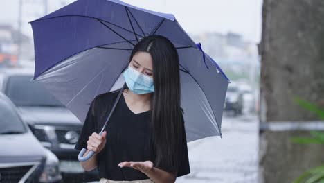 젊은 아시아 여성은 보호 얼굴 마스크를 착용하고 파란 우산을 들고 길가 에 서 있습니다. 샤워, 비가 오는 계절, 강우, 병에 걸릴 위험, 강한 강우, 호기심이 넘치는 손을 으십시오.