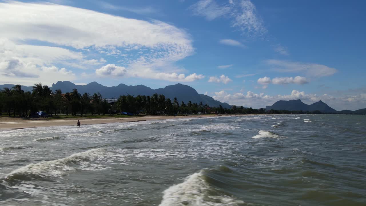 Metraje aéreo deslizante revelando la playa, silueta de personas caminando hacia el agua, cocoteros, montañas