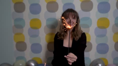 Young-Woman-Sitting-with-a-Sparkler-Against-Colorful-Background,-Surrounded-by-Balloons-on-the-Ground