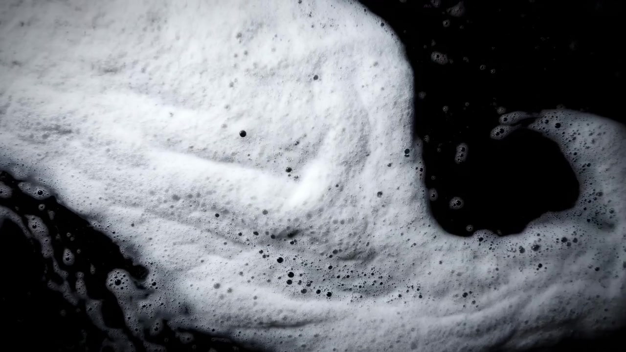 Primo piano macro che mostra una massa di schiuma bianca e bolle che si muove delicatamente su una superficie d'acqua scura. Una luce direzionale soffusa enfatizza la texture spumosa