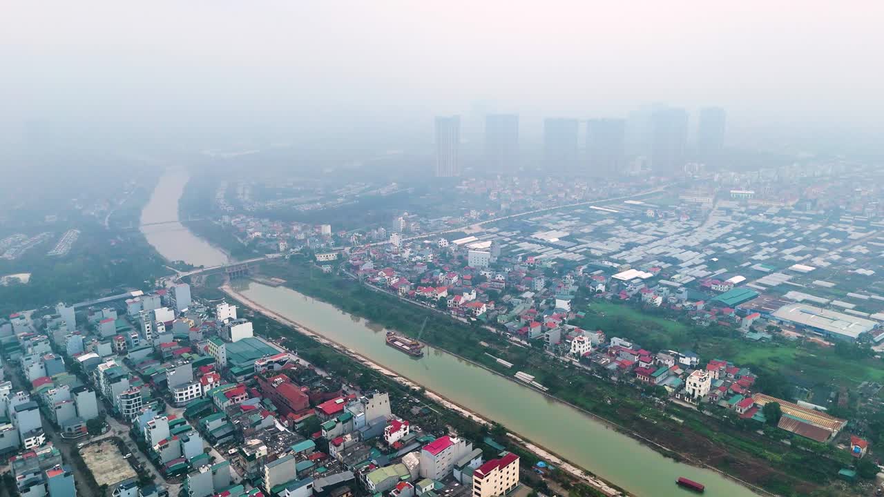 Aerial view of Bat Trang pottery village on the outskirts of Hanoi, Vietnam, with the river and residential buildings visible. The scene shows heavy pollution with smog over the area.