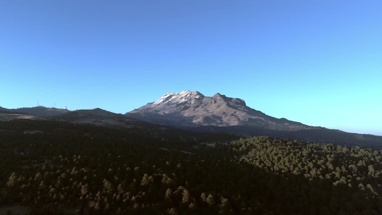 Ripresa aerea con drone di una montagna innevata che si erge sopra una fitta foresta, ampio paesaggio con cielo azzurro limpido, scenario naturale che mostra l'elevazione, la natura selvaggia e un ambiente tranquillo.