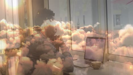 Animation-Eines-Geschäftsmannes,-Der-Im-Büro-Einen-Laptop-über-Sich-Bewegenden-Wolken-Benutzt