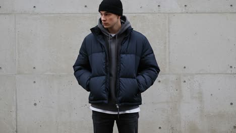 a young man in a stylish puffer jacket and beanie poses against a minimalist concrete wall, showcasing a trendy urban fashion look