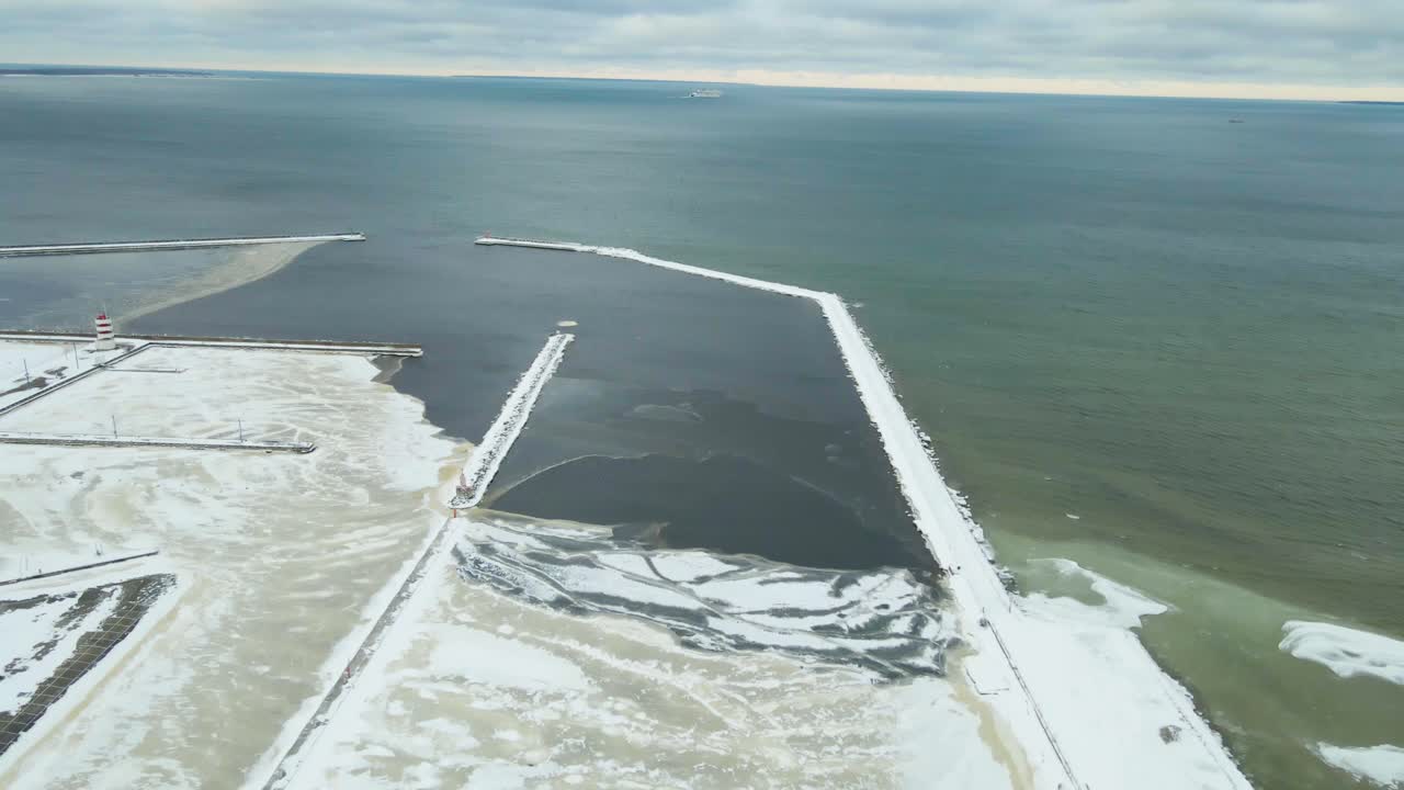Drone footage reveals a frozen harbor with snow covered piers, broken ice slabs, and cold Baltic water stretching toward the horizon under a cloudy winter sky