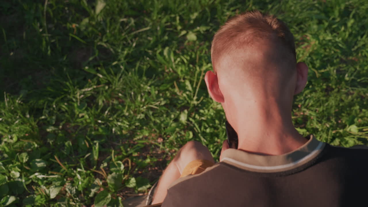 Young Man Examining Small Item While Seated Near Grass, Close Shoulder And Back Shots Show Focused Handling, Warm Sunlight And Leafy Background, Deliberate Careful Inspection And Slow Movements
