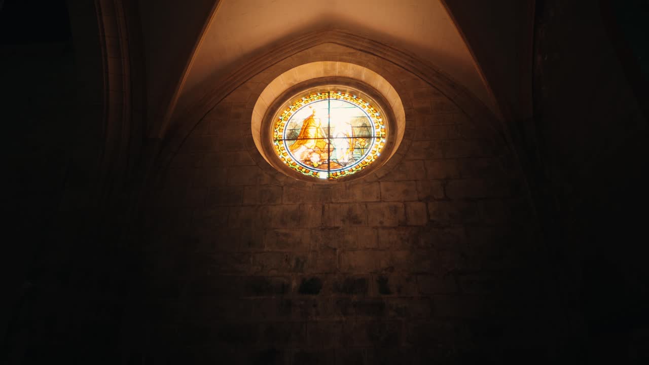 Um vitral circular representando uma figura religiosa ilumina um interior com arcos de pedra na vila de Issigeac, Dordogne. A luz baixa realça a textura da alvenaria.