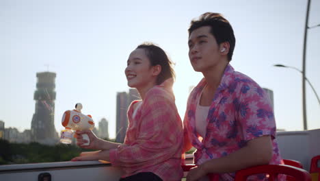 couple of asiatic lovers travelling on open air bus with skyline cityscape at distance during sunset playing with a bubble soap toy gun