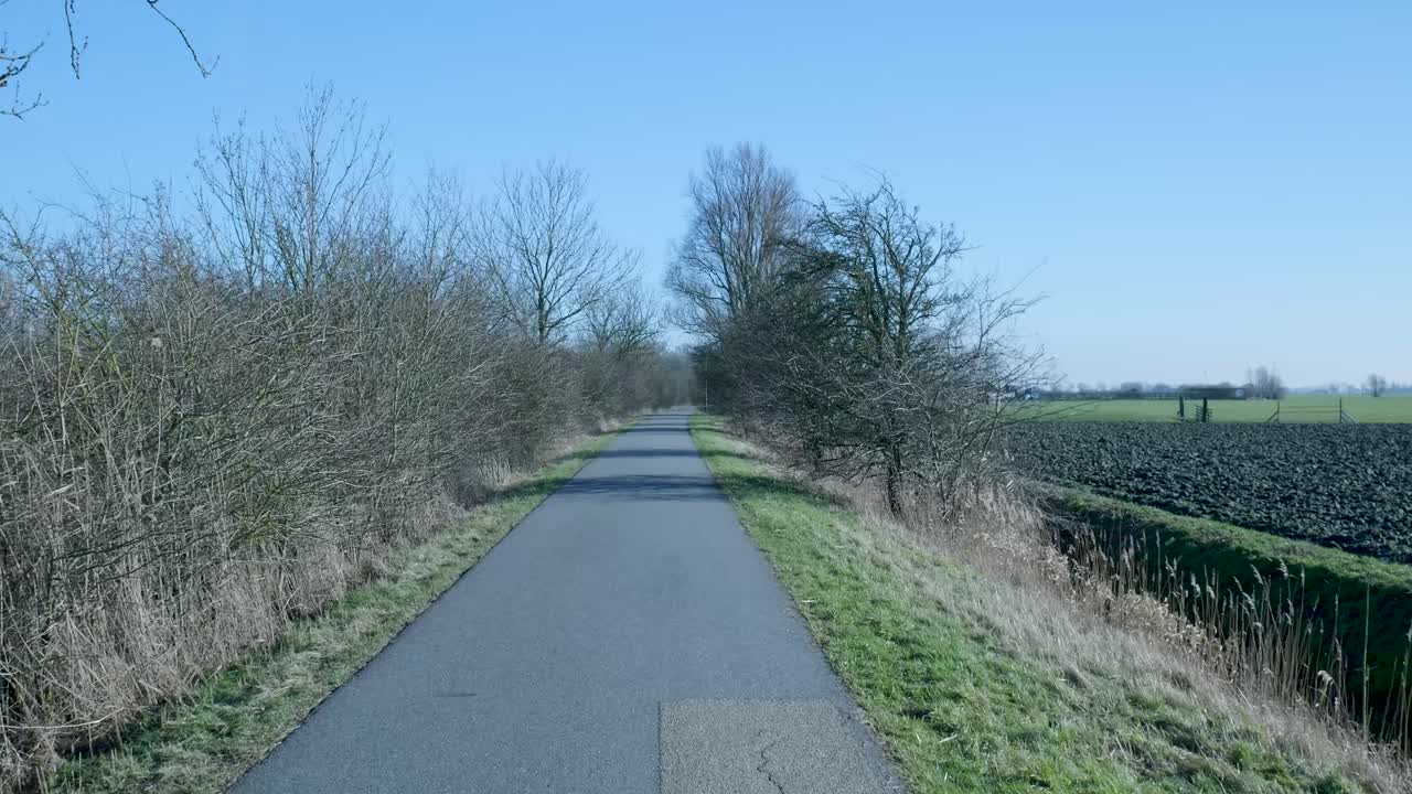 Førstepersonsbillede af en landlig sti omkranset af nøgne træer og marker