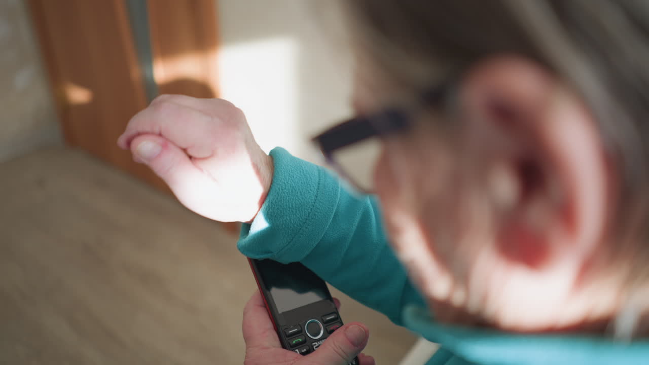 Elderly woman with glasses, wearing turquoise fleece sweater, looking at her mobile phone, adjusting her glasses with one hand. Soft light coming from behind her, focusing on her hands