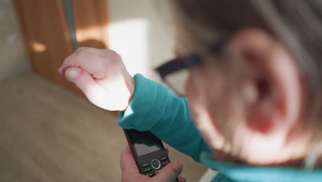 elderly woman with glasses, wearing turquoise fleece sweater, looking at her mobile phone, adjusting her glasses with one hand. soft light coming from behind her, focusing on her hands