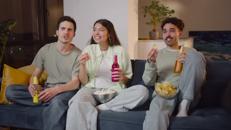 three friends watching comic movie on television sitting on couch, eating popcorn and chips and drinking soda