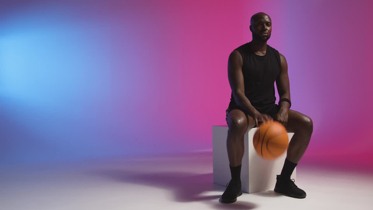 retrato de estudio de un hombre sentado rebotando una pelota de baloncesto contra un fondo iluminado de rosa y azul