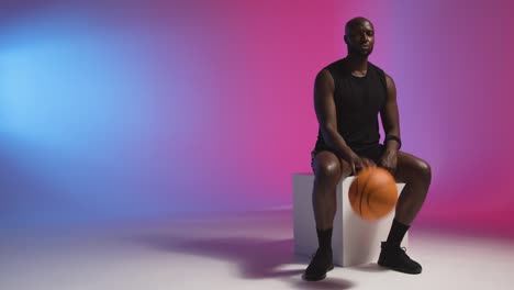 studio portrait shot of seated male basketball bouncing ball against pink and blue lit background