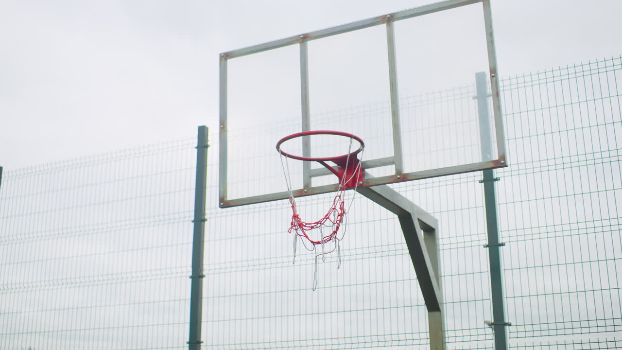 Canestro da basket all'aperto con una palla che descrive un arco verso il canestro attraverso la rete su un campo recintato vuoto sotto un cielo nuvoloso. Il canestro in metallo rosso e il tabellone trasparente usurato rivelano un ambiente da parco giochi urbano, traiettoria al rallentatore