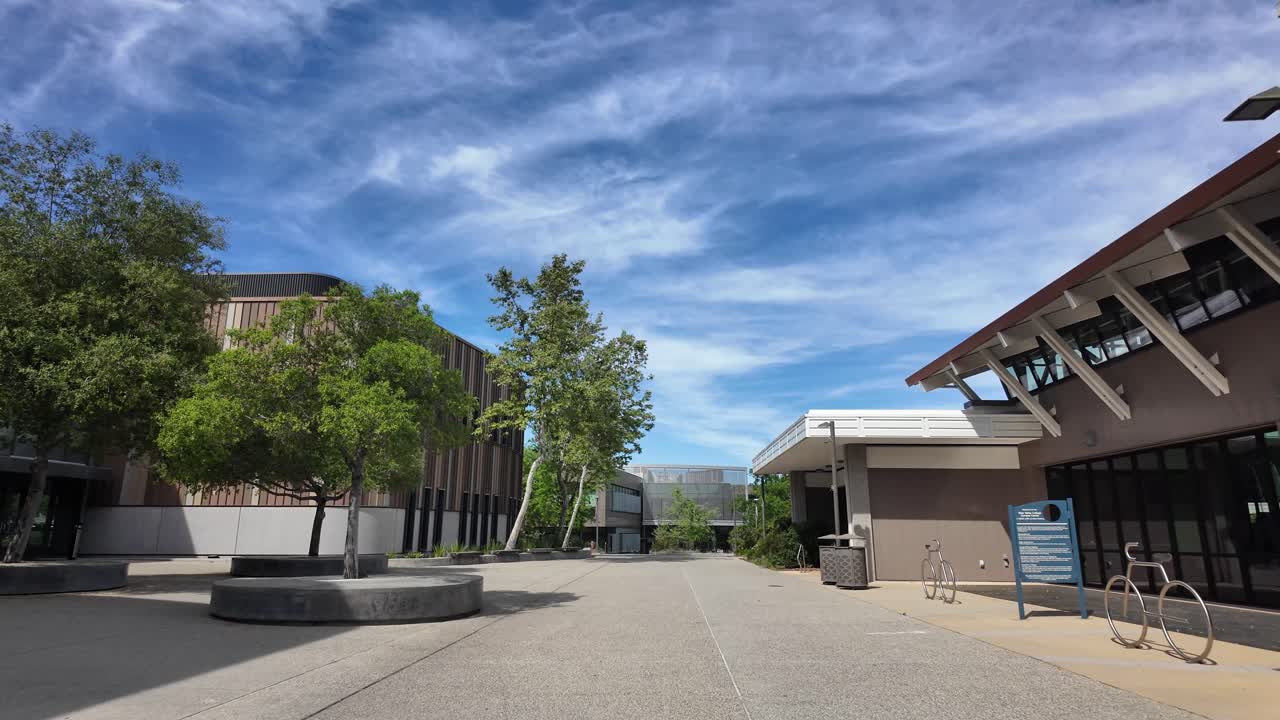 Ripresa in soggettiva mentre si cammina attraverso il campus moderno e deserto del West Valley College a Saratoga, California, che mostra architettura contemporanea e cortili paesaggistici sotto un cielo blu.