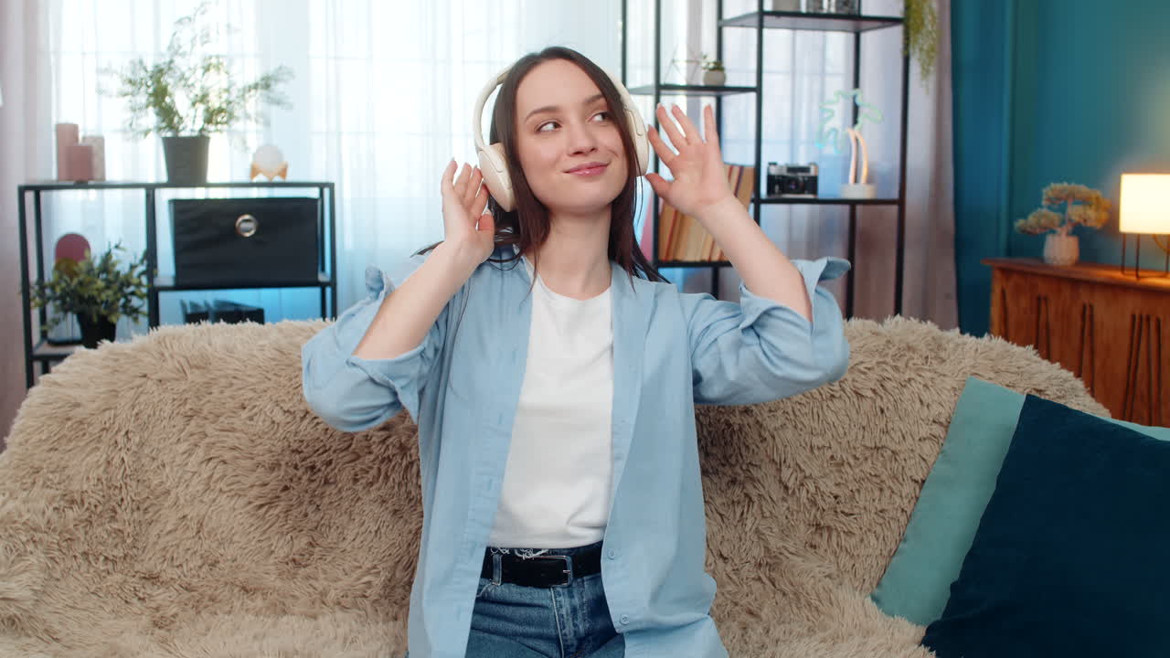 Young woman wearing wireless headphones on sofa dancing smiling happily enjoying favorite song