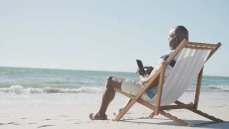 Senior-african-american-man-on-deck-chair,-using-smartphone-at-beach,-copy-space,-in-slow-motion