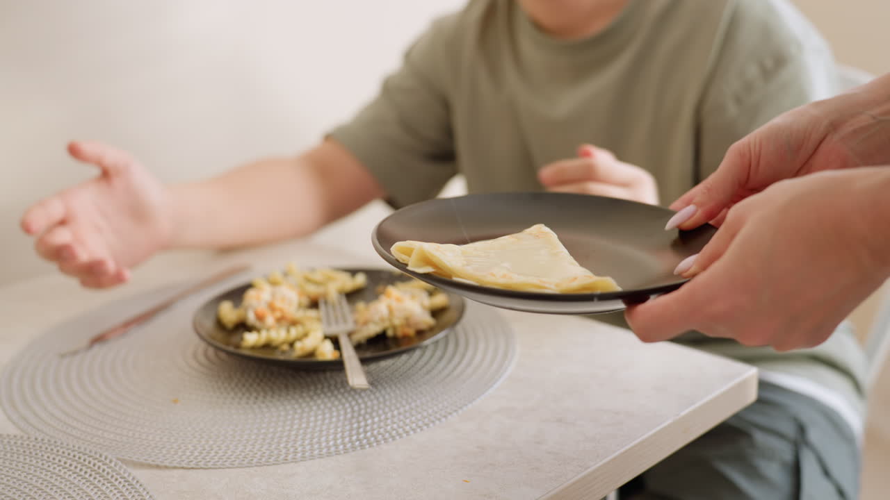 Close up of woman in green shirt carrying egg on black plate in kitchen, shifting dish toward boy seated off screen, hands near stove, cooking moment showing care