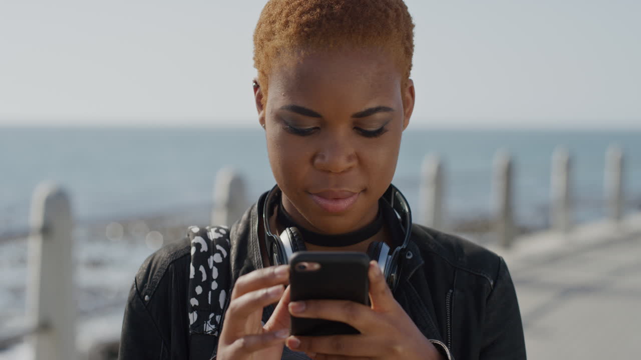 ritratto bella giovane donna afroamericana che usa lo smartphone messaggistica navigazione online invio messaggi in attesa sul mare soleggiato sorridendo felice indossando moda elegante