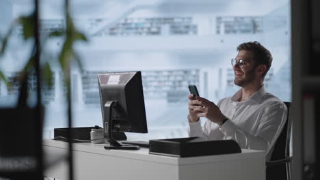 Smiling-young-guy-using-mobile-apps-sit-at-table.-Male-millennial-professional-holding-modern-smartphone-texting-message-in-office.-Young-businessman-using-helpful-mobile-apps-for-business-time