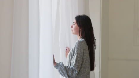 pensive woman standing looking outside through a window at home in the morning