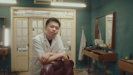 young asian barber leaning on chair posing on camera
