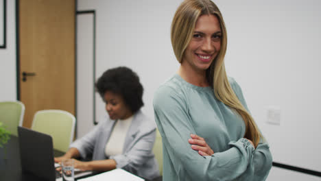 Retrato-De-Una-Mujer-De-Negocios-Caucásica-Sonriendo-En-La-Oficina,-Con-Un-Colega-Trabajando-En-Segundo-Plano