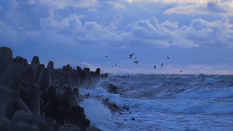 Große-Stürmische-Wellen,-Die-Sich-Gegen-Die-Wellenbrecher-Des-Nördlichen-Piers-Bei-Liepaja-Brechen,-Bewölkter-Abend,-Möwen,-Die-Im-Starken-Wind-Treiben,-Zeitlupenweitwinkelaufnahme