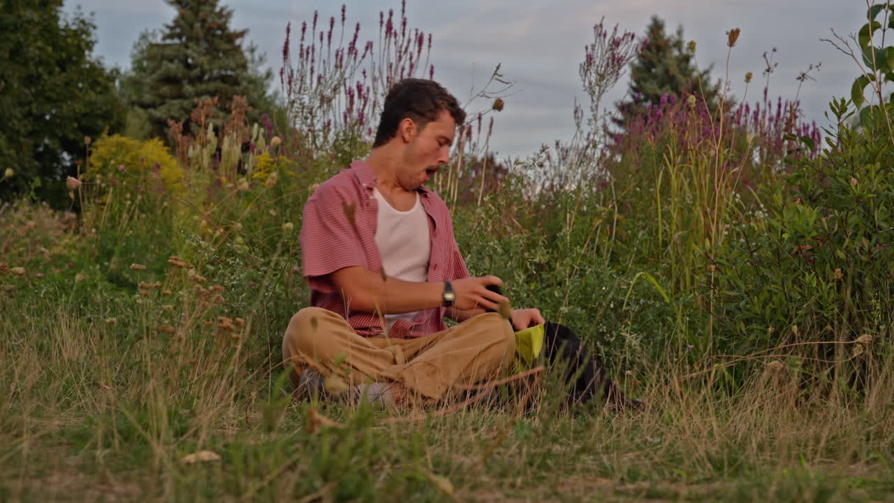 A young Gen Z man sits in a wild meadow, checks his phone, becomes tired, then disconnects to appreciate the cloudy colorful sky and meditate in nature. Perfect for digital detox themes
