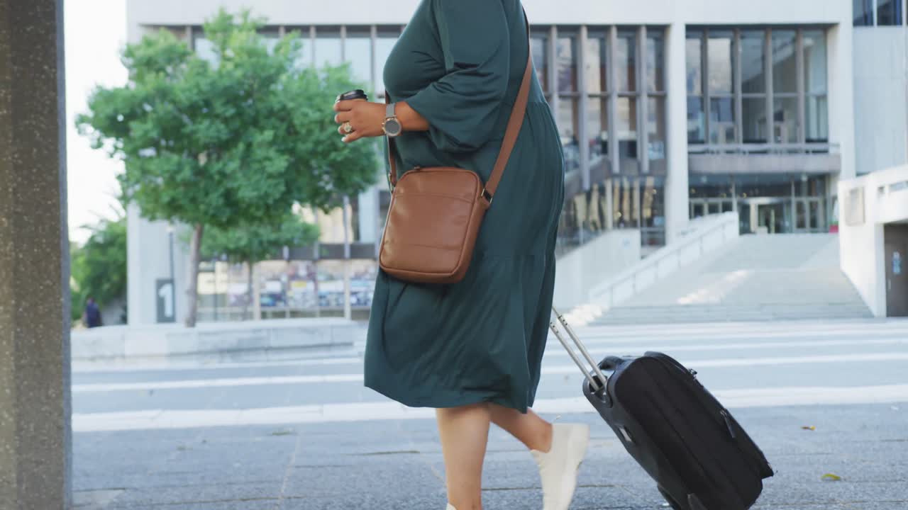Plus size biracial woman walking with luggage and takeaway coffee in city
