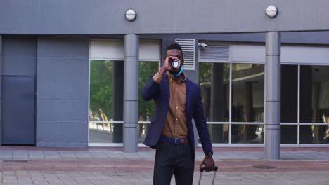 african american businessman with face mask walking drinking coffee in street