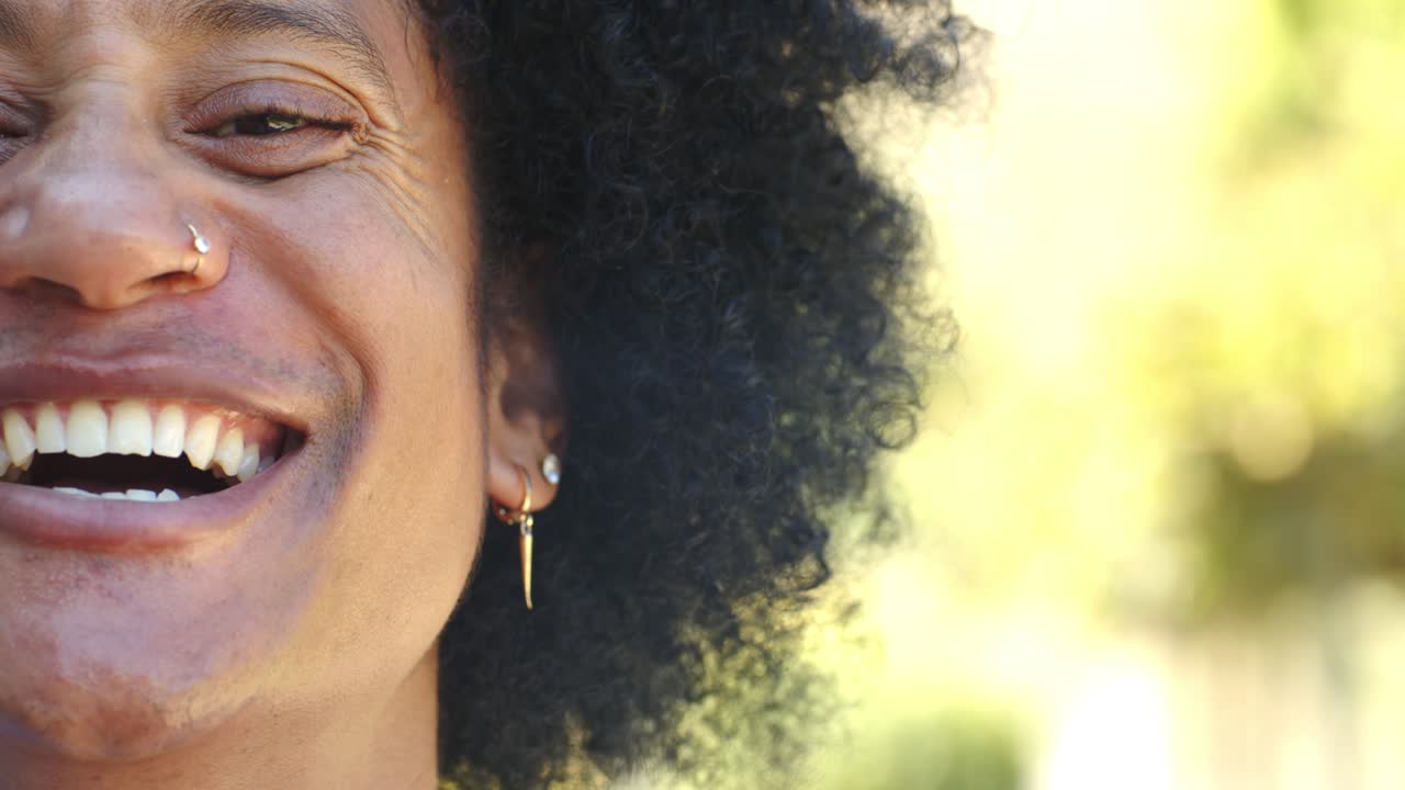 African American adult smiling, laughing, leaning on sunlit street with nose hoop, ear studs, teeth