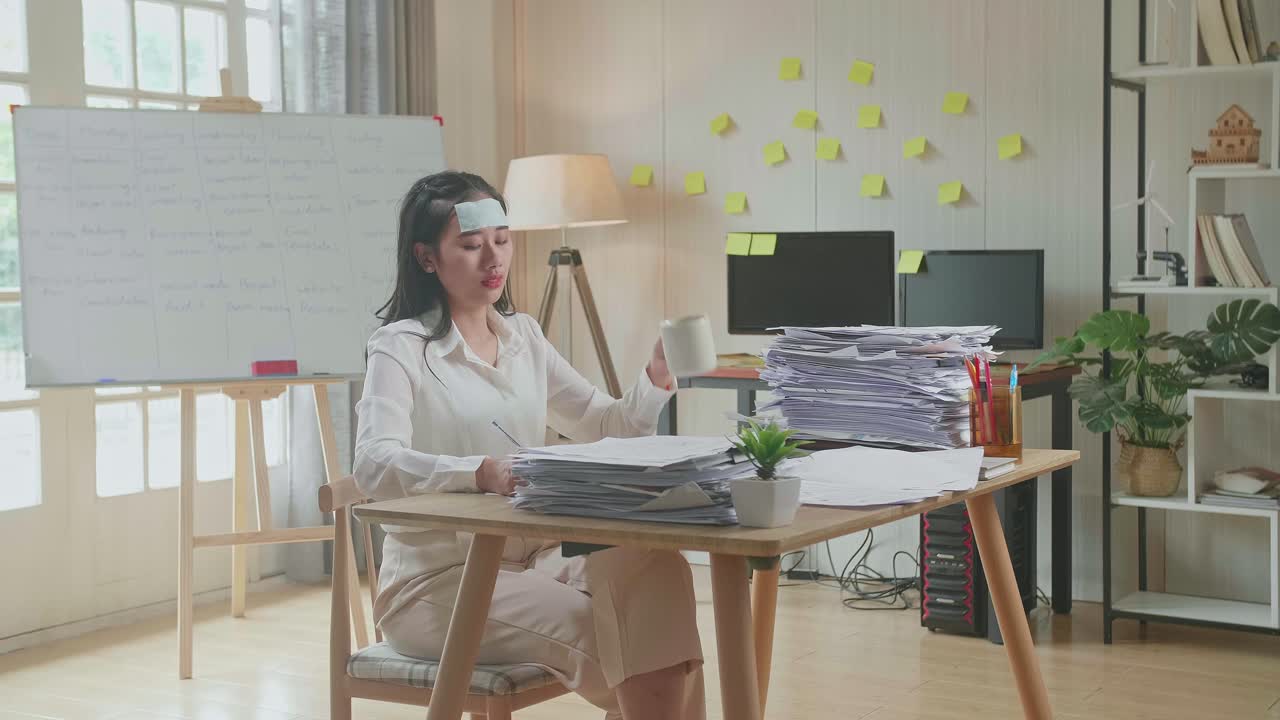 병든 아시아 여성이 사무실에서 문서와 함께 열심히 일하면서 물을 마시고 있습니다.