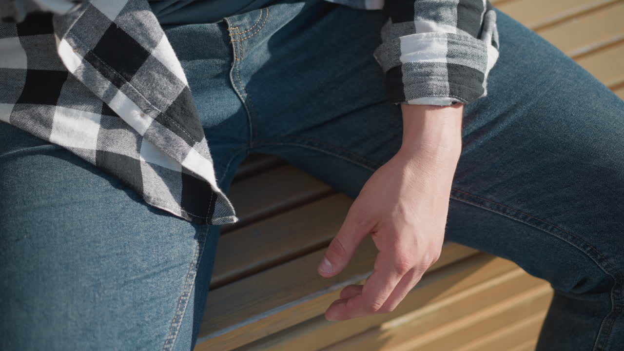 partial view of man in checkered shirt and jeans sitting on wooden bench outside during sunny day with hand resting beside leg after taking red pill medicine in quiet peaceful environment