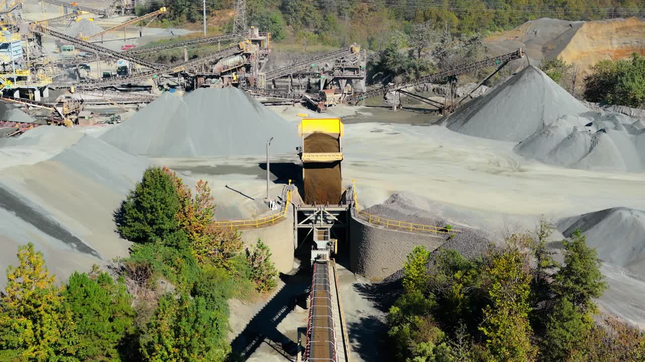 Dump truck unloads fill near gravel mound overlooking hopper and conveyor belt processing plant, aerial pullback