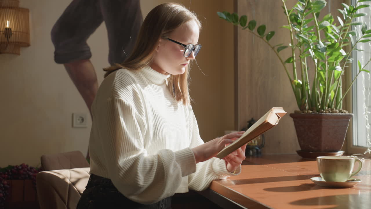 Young lady in white sweater reading book near window as soft sunlight highlights her focused expression, cozy indoor setting with warm tones, potted plant, and coffee cup placed on wooden table