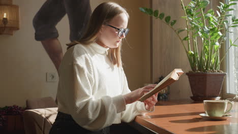 young lady in white sweater reading book near window as soft sunlight highlights her focused expression, cozy indoor setting with warm tones, potted plant, and coffee cup placed on wooden table