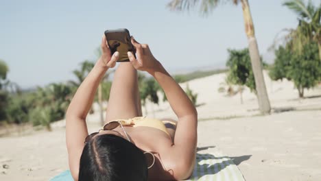 Hispanische-Frau-Mit-Sonnenbrille,-Die-Auf-Dem-Rücken-Am-Strand-Liegt-Und-Ihr-Smartphone-Benutzt,-Zeitlupe