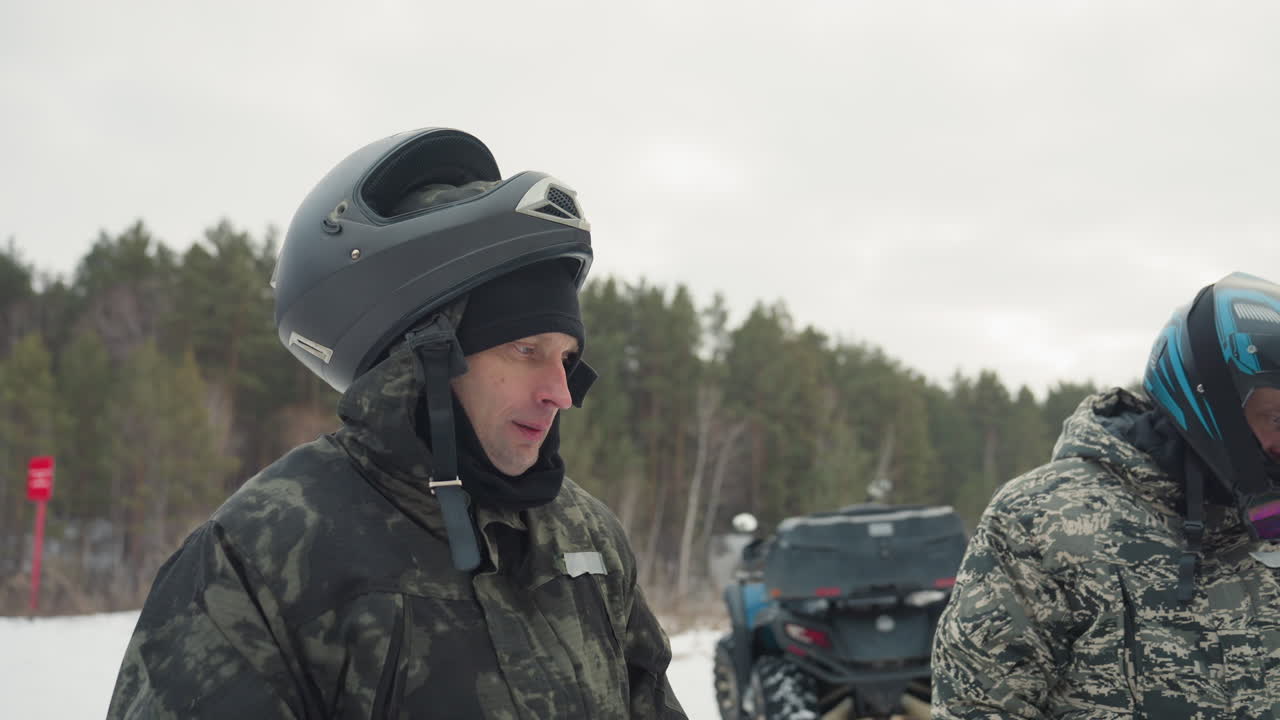 Pilota ispeziona il casco vicino a un quad in una radura innevata, primo piano di profilo di un uomo concentrato con giacca mimetica e berretto di lana sotto il casco, respiro ghiacciato e sguardo intento, quad parcheggiato dietro, pino