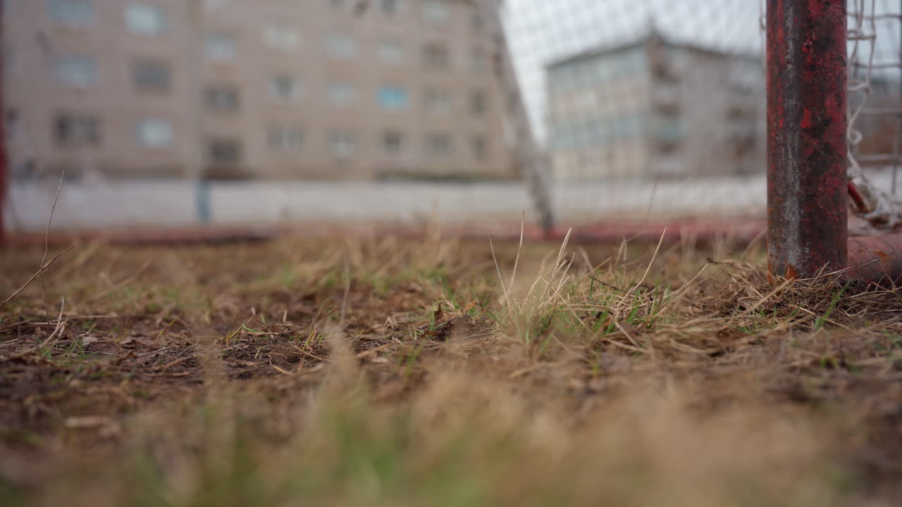 Balón de fútbol que se acerca desde un ángulo bajo hacia la portería, trayectoria y efecto visibles sobre hierba seca, terreno urbano con edificios de apartamentos difuminados al fondo, movimiento dinámico y crudeza desde la base