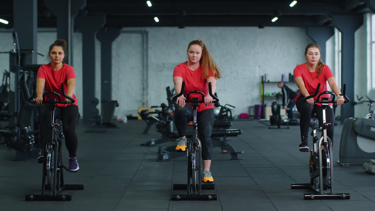 건강한 백인 여성 그룹이 체육관에서 정지 자전거 기계 자전거에서 운동을합니다.
