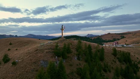 Aerial-orbit-shot-of-waving-Romanian-flag-and-cross-on-top-of-Dichiu-Mountain-at-sunset-in-Romania