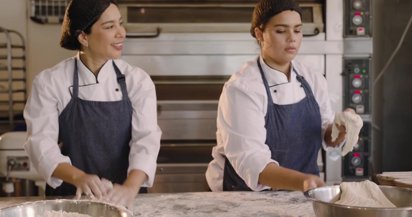 Diverse female bakers scraping dough from bowl, cutting rounds on dusty bench into baskets to proof