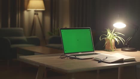 laptop computer showing green chroma key screen stands on a desk in the living room. in the background cozy living room in the evening with warm lights on