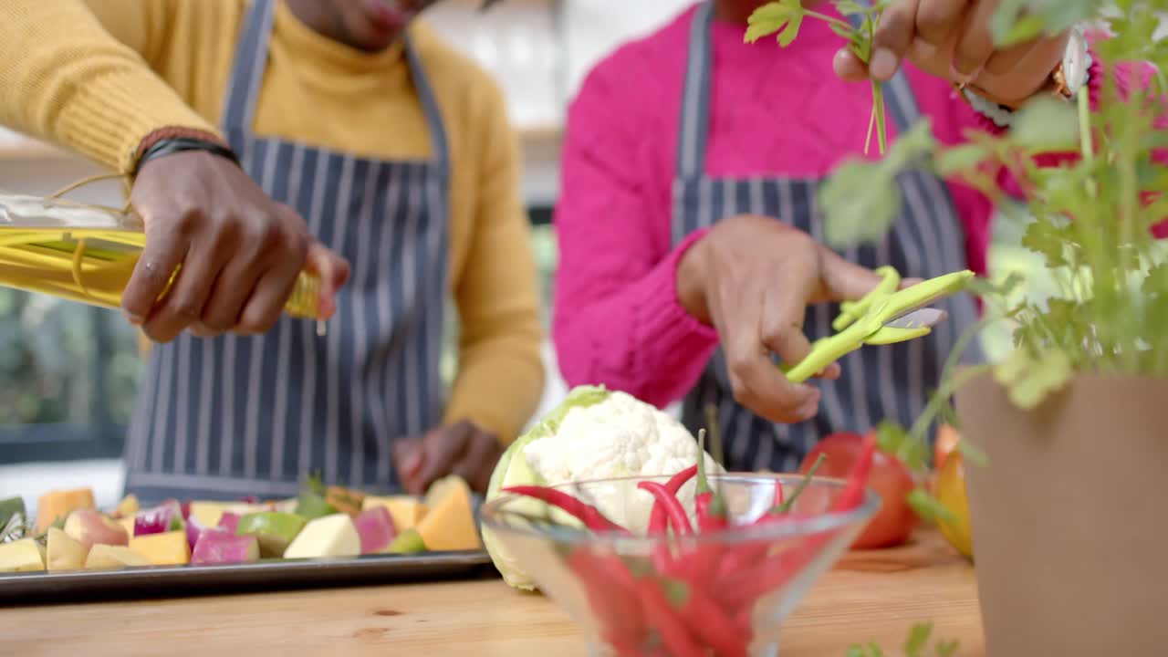 Plano médio de casal afro-americano de aventais derramando azeite nos vegetais na cozinha, em câmera lenta.
