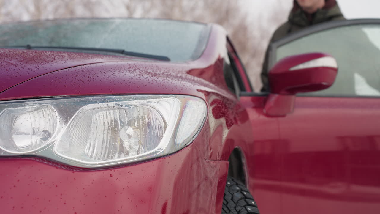 Front angle view of red car with water droplets on headlight and hood during winter as young man in winter jacket enters vehicle, snowy background with bare trees enhances cold season atmosphere